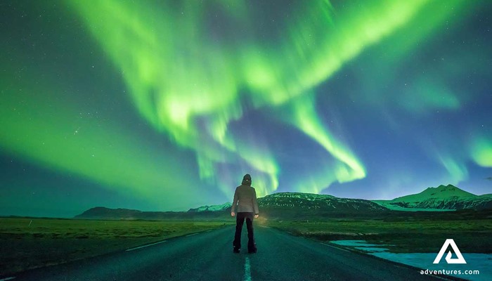 man standing on a road below northern lights in iceland