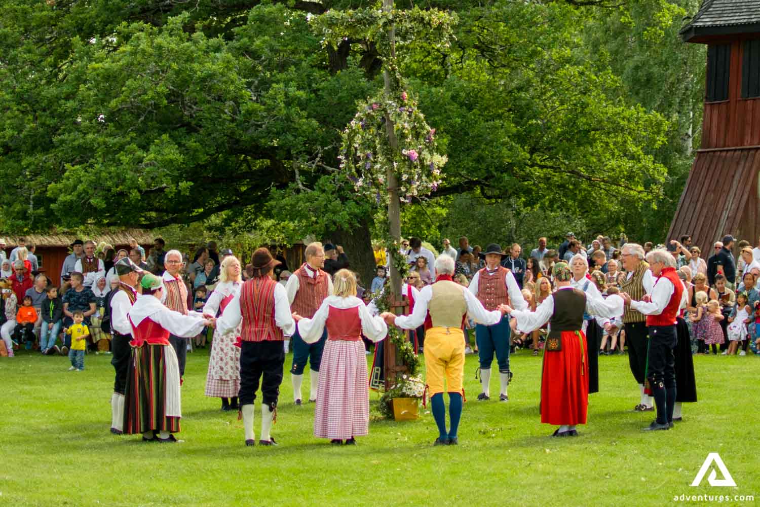 Foto de evento midsommar na suécia. Sobre um vale verde, pessoas dançam de mãos dadas ao redor de uma figura feita de grama