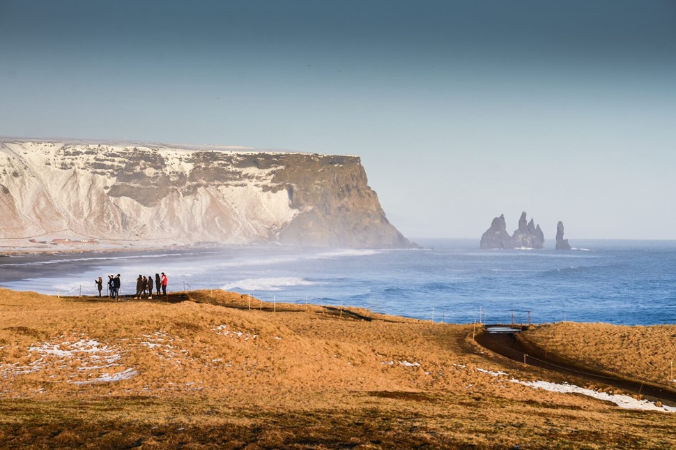 People on a coastal overlook with Reynisdrangar sea stacks and snowy cliffs in the background in Iceland.
