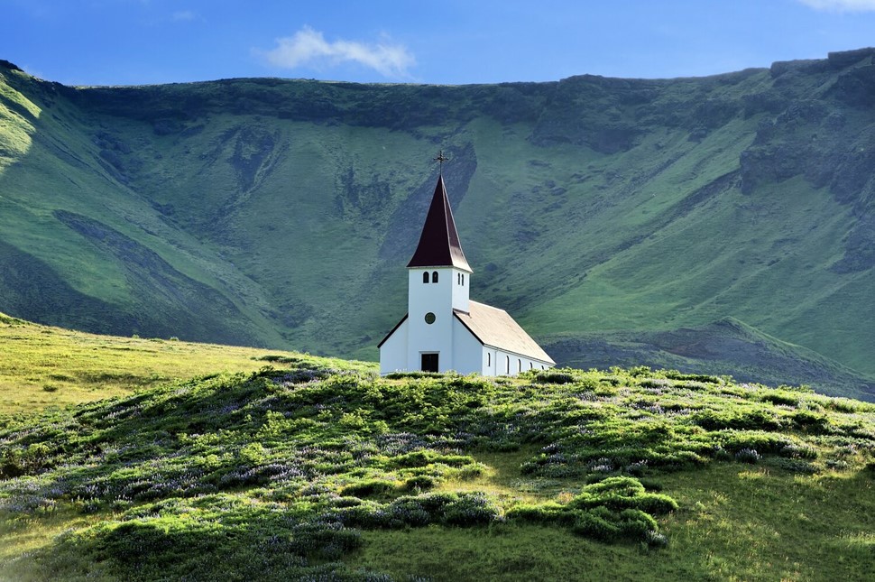 Church with a red pointed steeple in lush green hills under a bright blue sky in Vik Iceland.