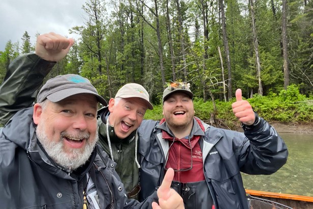 Three happy fisherman showing thumbs up, standing on boat in river, behind  them a background evergreen forest 