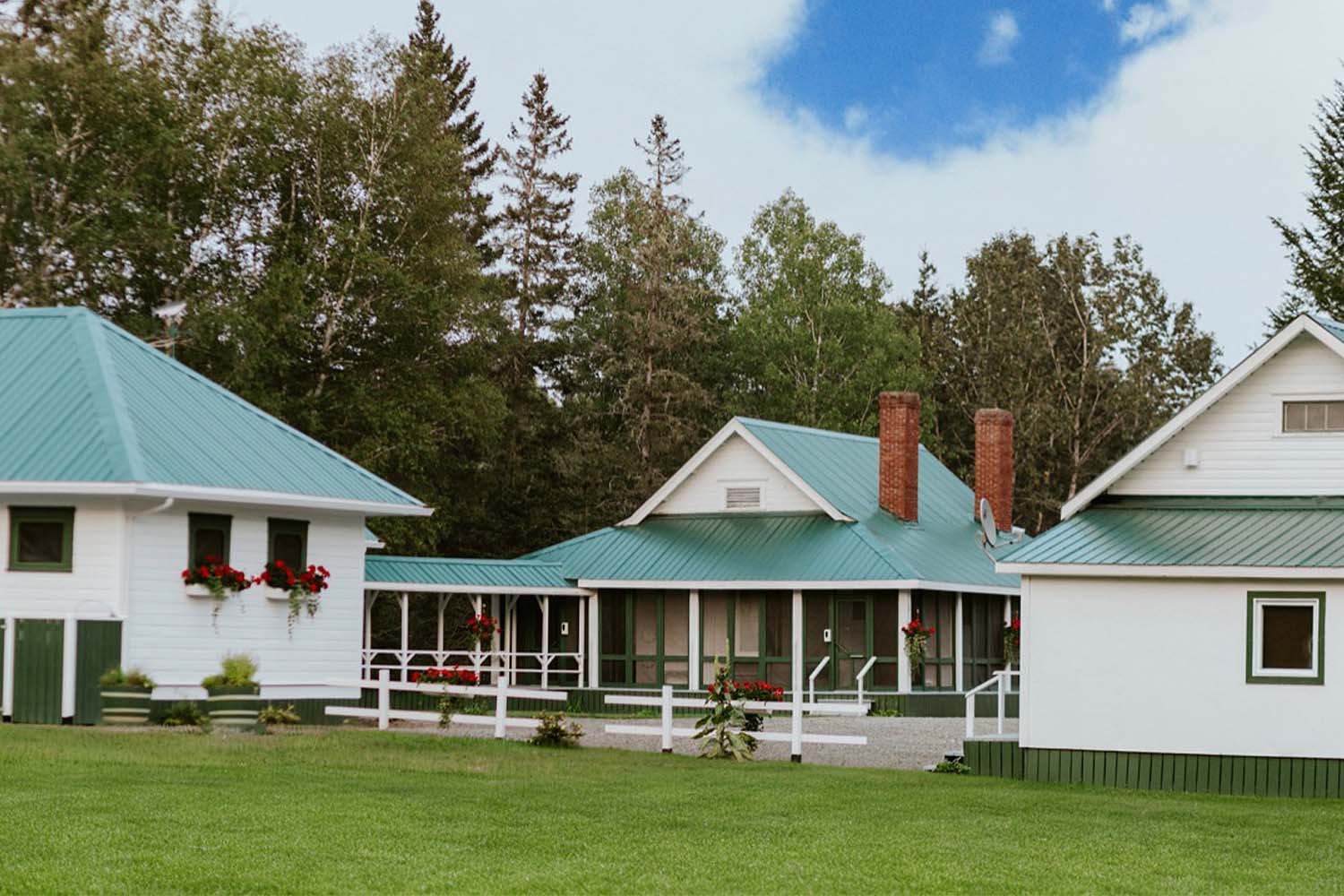 White wooden fishing accommodation building with green roof and hanging red flowers with evergreen trees in background and blue sky 