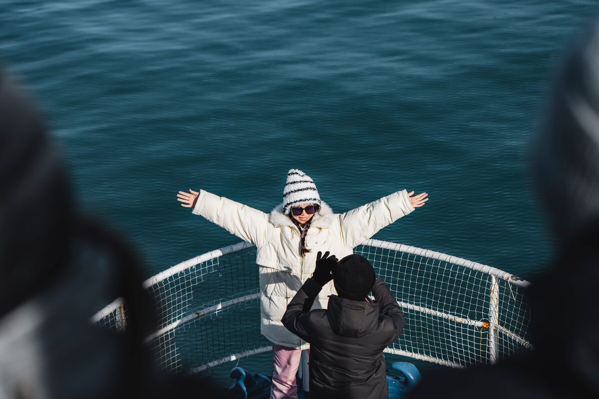 Female dressed in white coat with arms up in air posing for photo on boat.