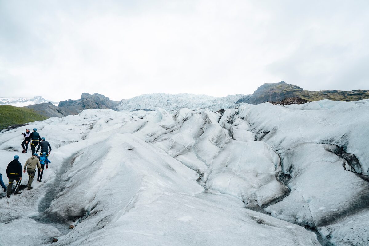 Distant view of a small tour group climbing up the Skaftafell glacier, surrounded by expansive ice fields with green mountains in background.