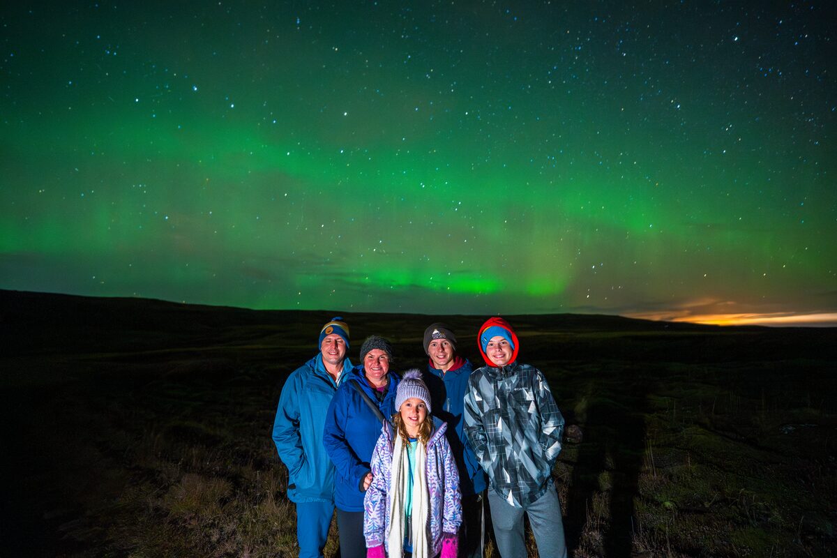 Family posing for photograph in late evening standing underneath starry sky with northern lights.