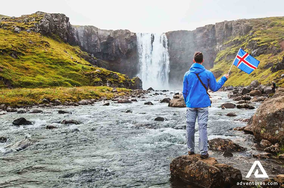 a man with an icelandic flag near a small waterfall